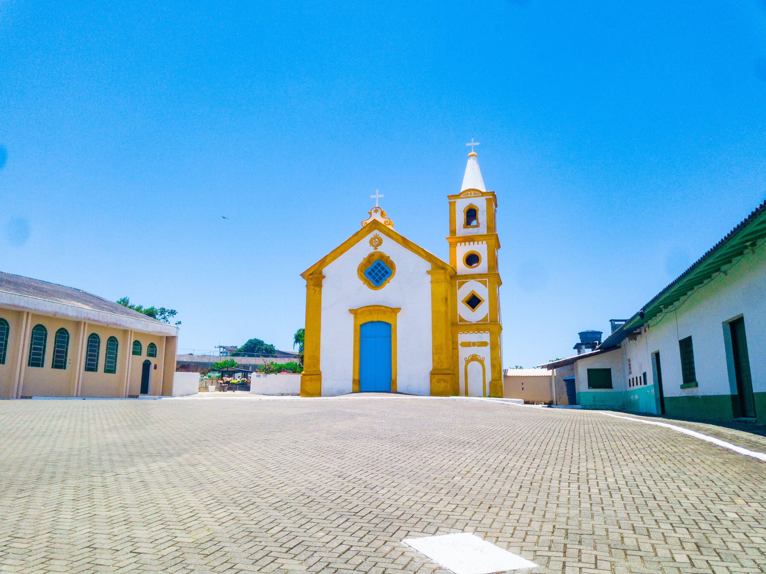 Fachada da Capela São João Batista, em branco e amarelo, com portão azul e torre sineira