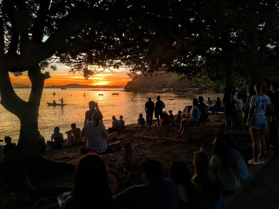 Pessoas reunidas à beira-mar assistindo ao pôr do sol na Praia do Cascalho, em Penha