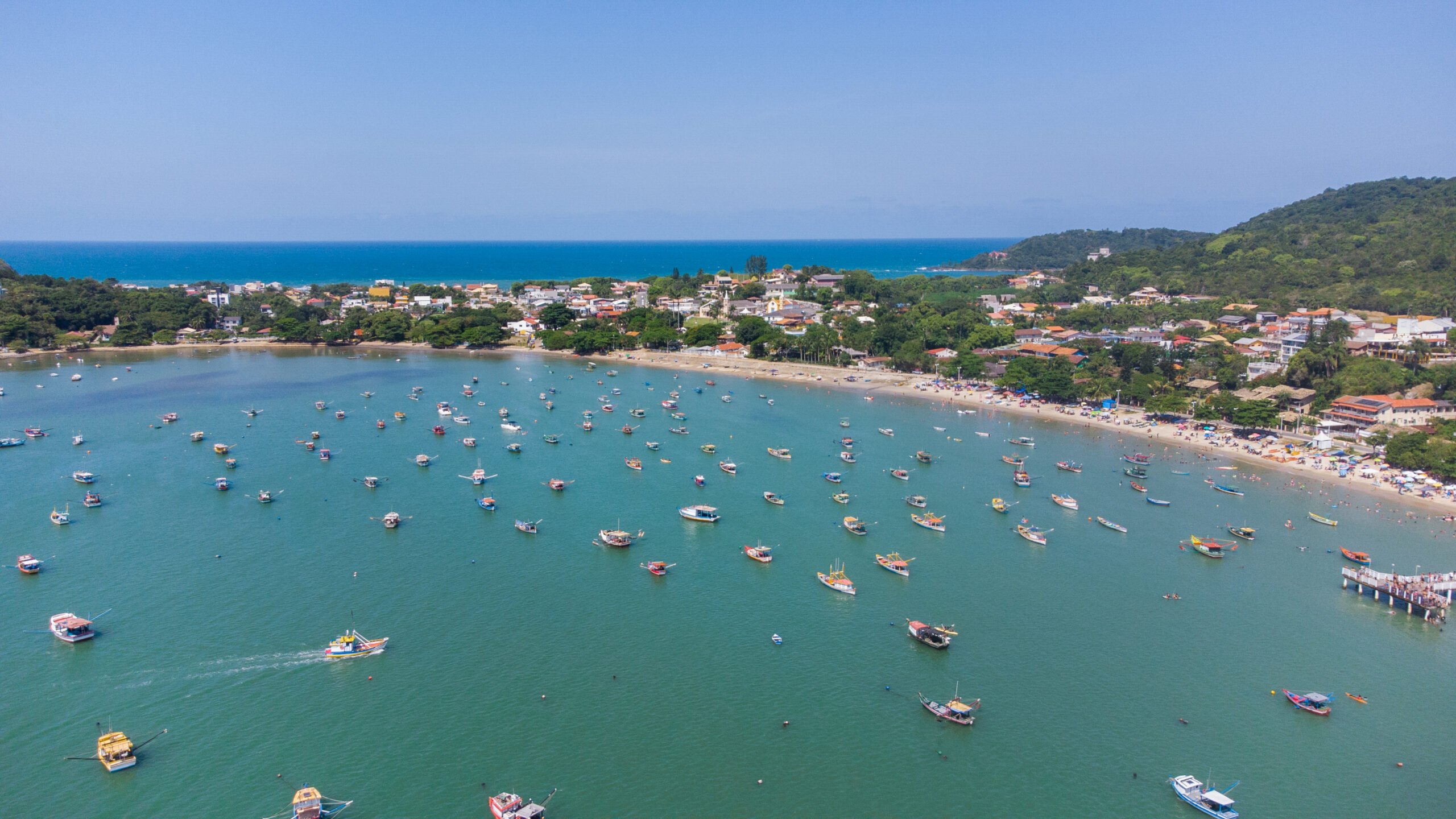 Vista aérea da Praia da Armação, em Penha, com a enseada repleta de barcos de pesca e o trapiche à direita