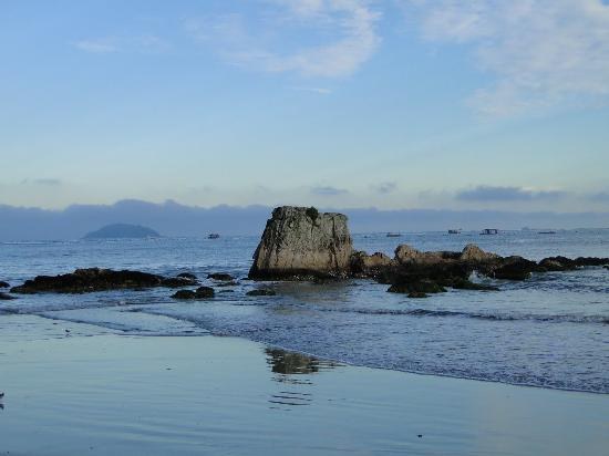 Praia da Fortaleza, em Penha, ao amanhecer, com uma grande pedra emergindo do mar calmo e ilha ao horizonte